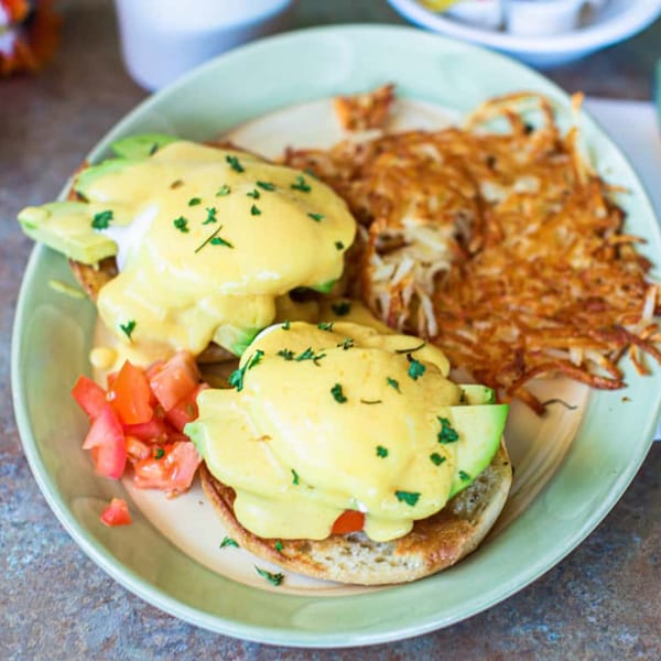 Lucky's avocado eggs Benedict with hash browns and diced tomatoes on a light green plate.