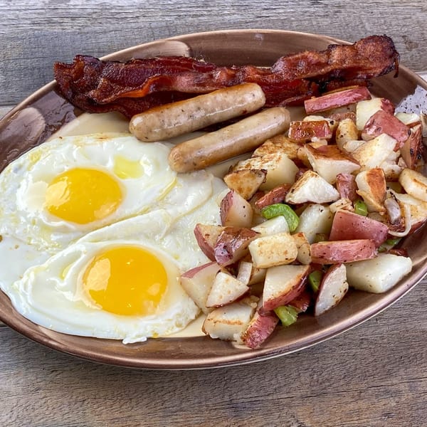 A breakfast plate with two fried eggs, two sausages, crispy bacon, and roasted potatoes.
