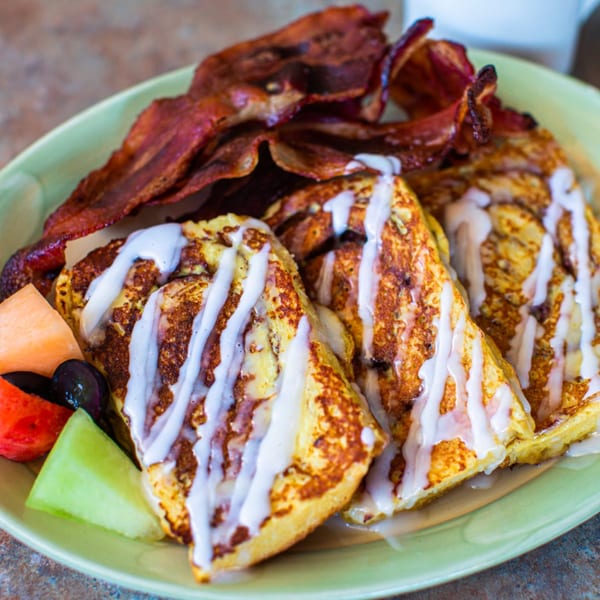 French toast with white icing drizzle, crispy bacon, and a side of fresh fruit on a green plate.