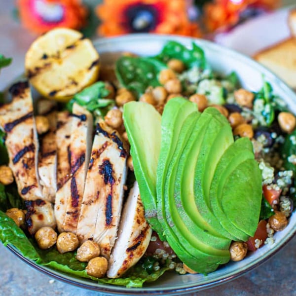 Grilled chicken and avocado slices on a salad with chickpeas, quinoa, and a grilled lemon.